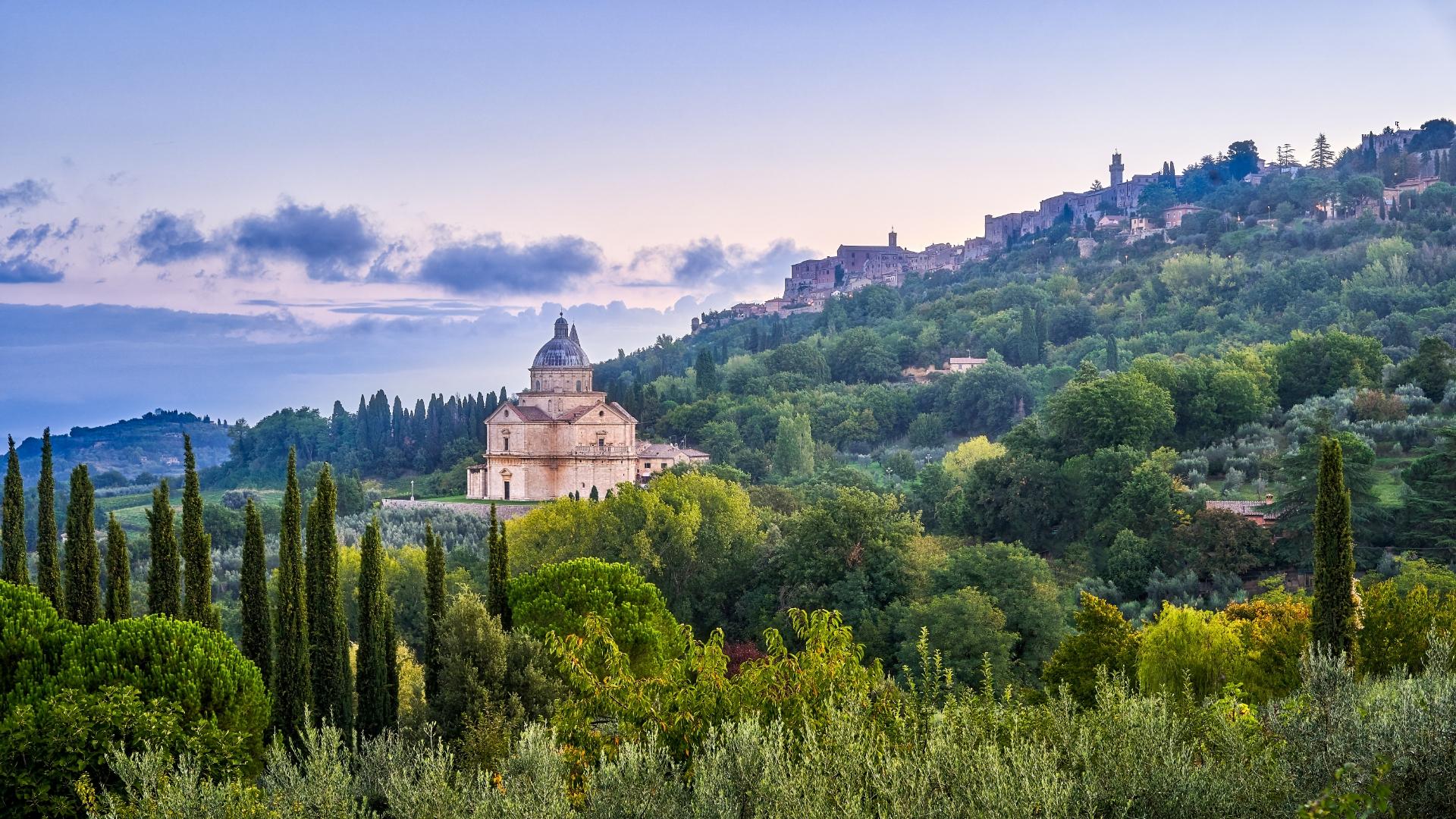Paesaggio collinare toscano, chiesa storica tra cipressi e verde lussureggiante al tramonto.