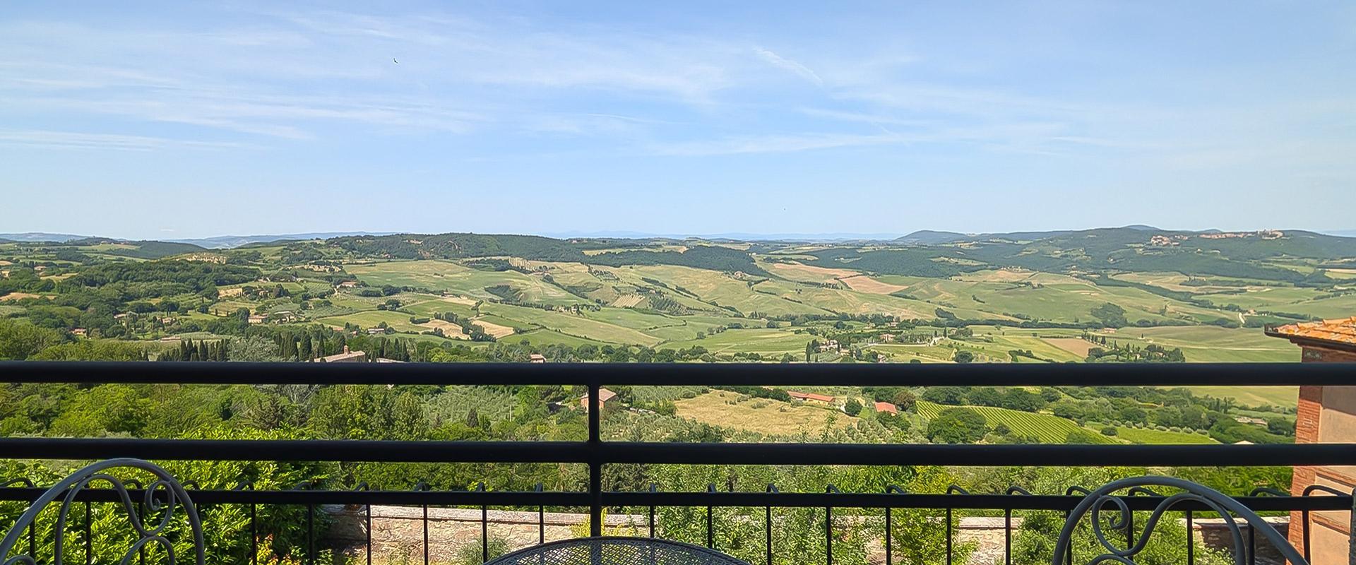 Vista panoramica su colline verdi, cielo azzurro e un terrazzo con tavolo in ferro battuto.