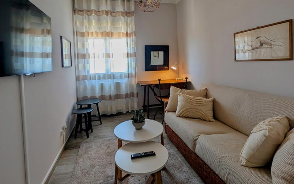 Soggiorno accogliente con divano, tavolini, lampada moderna e luce calda che filtra dalle tende.