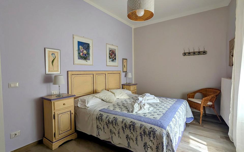 Camera accogliente con letto sfatto, pareti lilla e luce naturale che entra dalla finestra.